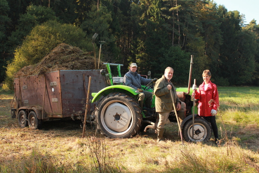 Ein Mann und eine Frau stehen vor einem Traktor. Sie halten Heugabeln in der Hand. Auf dem Traktor sitzt noch ein Mann. Am Traktor hängt ein Wagen voll mit Gras.